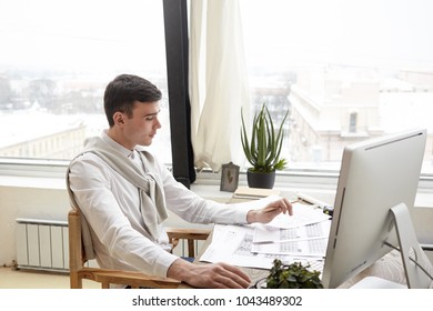 Serious Clean Shaven Man Architect Dressed Formally Working With Blueprints In His Office, Sitting In Front Of Computer. Concentrated Interior Designer Studying Layout Of Housing Project On Desk