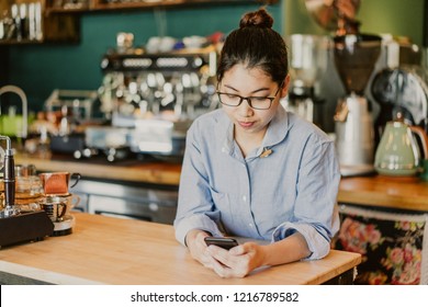 Serious Coffee Shop Owner Reading Message On Phone While Waiting For Clients. Busy Young Asian Female Entrepreneur Using Smartphone At Workplace. Modern Small Business Concept