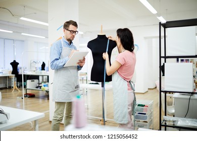 Serious colleagues taking measurements from mannequin while working on new garment in studio: female assistant measuring mannequin while tailor writing out it in sketchpad - Shutterstock ID 1215056452