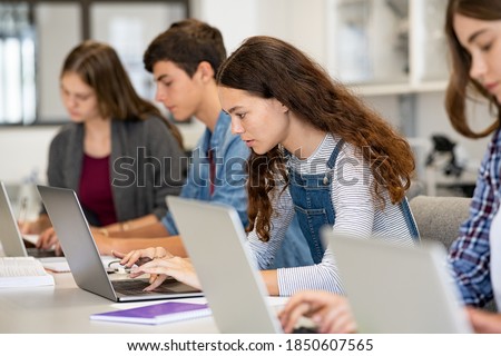 Serious college students studying on laptop sitting in a row in library. Young university multiethnic students using computer for study in classroom. Side view of girl typing on laptop during lesson.