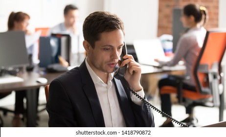 Serious Company Employee Male In Formal Suit Sitting In Shared Office Room Coworking Space With Other Workers Talking On Landline Phone With Client. Call Centre Support Distance Communication Concept