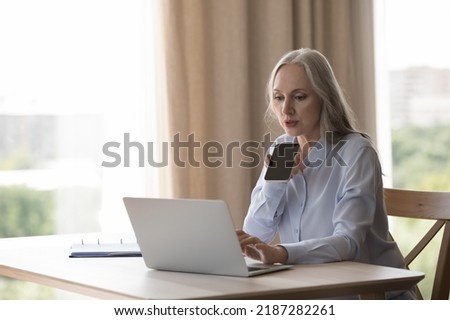 Serious concentrated elderly business woman recording voice message, giving command to virtual assistant, using online virtual app on laptop, talking to client on mobile phone
