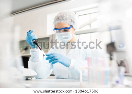 Serious concentrated female microbiologist in sterile clothing and safety goggles sitting at table and dropping reagent in petri dish while doing research in laboratory