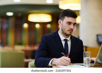 Serious Concentrated Handsome Business Expert In Suit Sitting At Table In Restaurant And Editing Information In Papers While Using Laptop