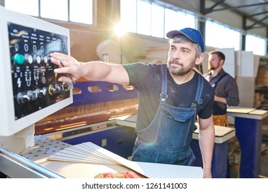 Serious concentrated handsome gray-haired workman in cap and denim overall pushing button on control panel while setting up factory machine in industrial shop - Shutterstock ID 1261141003