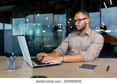 Serious and concentrated hispanic programmer working inside office, man in shirt writing codes for software using laptop.
