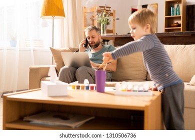 Serious concentrated modern father being too busy sitting on sofa and calling on phone while using laptop in living room, little son choosing paintbrush