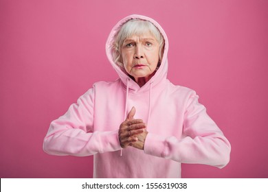 Serious concentrated old woman stand alone in fight position. Look straight on camera. Wewar pink hoody. Hold one hand in fist and touch it to another. Isolated over pink background