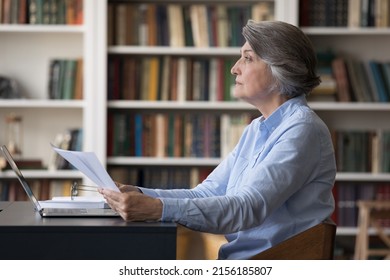 Serious Concentrated Older Business Woman Analyzing Paper Documents At Workplace, Using Laptop In Library, Looking Away, Thinking Over Agreement Terms, Feeling Doubt, Making Decision