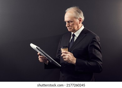 Serious Concentrated Senior Executive In Black Suit Standing Against Isolated Background And Drinking Takeout Coffee While Working With Papers