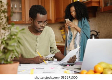 Serious And Concentrated Young African-American Male Wearing Spectacles Paying Bills Online, Using Banking App On His Mobile Phone. People, Paperwork, Finances, Family Budget And Economy Concept