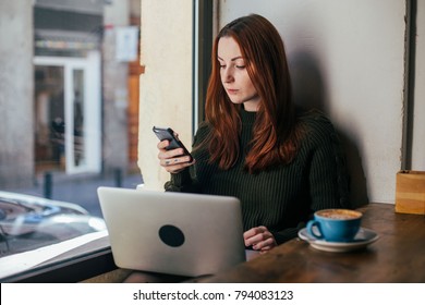 Serious And Concentrated Young Businesswoman Or Student Freelancer Works In Coworking Hub Or Coffee Shop With Laptop And Smartphone, Scrolls Through News Feed On Social Media,