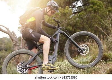 Serious And Concentrated Young European Cyclist Wearing Black Cycling Clothing, Helmet, Eyeglasses And Gloves Switching Speed Mode On His Pedal-assist Electric Bike Before Riding Uphill. Flare Sun