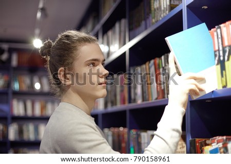 Serious concentrated young European man with hair bun taking book from bookshelf in bookstore while searching for popular bestseller. 20 year old male choosing detective to read in his spare time