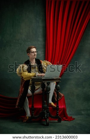 Serious, concentrated young man working on laptop against dark green, vintage background. Male model as a duke, prince, royal person. Concept of comparison of eras, history, renaissance art, remake