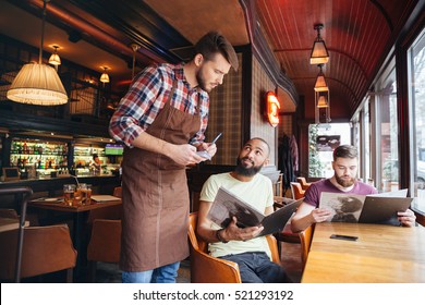 Un jeune garçon sérieux et concentré debout sur ordre de deux beaux hommes barbus au café : photo de stock