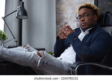 Serious Confident African Businessman In Ripped Jeans Sits With His Legs Folded On The Table And Looks At The Camera. Concept Of A Free Lifestyle Of An Entrepreneur Or Freelancer.