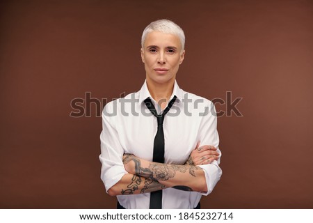 Serious and confident blond businesswoman in formalwear crossing arms with tatoos by chest while standing in front of camera in isolation