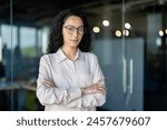 Serious and confident business woman inside office at workplace, portrait of a Latin American boss. A woman with curly hair and wearing glasses looks confidently at the camera with her arms crossed.