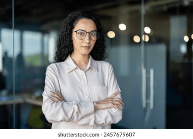 Serious and confident business woman inside office at workplace, portrait of a Latin American boss. A woman with curly hair and wearing glasses looks confidently at the camera with her arms crossed. - Powered by Shutterstock