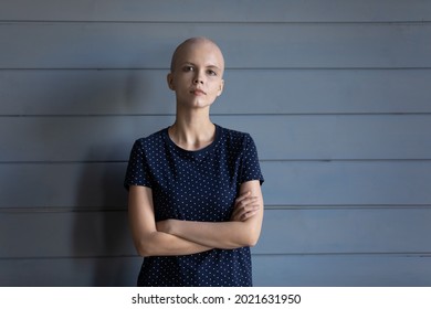 Serious confident cancer survivor head shot portrait. Young hairless woman with arms folded looking at camera, standing against studio background. Patient fighting for life against oncology disease