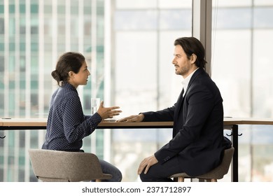 Serious confident diverse colleagues discussing project strategy, cooperation in co-working space. Young female Indian employee talking to male boss, company business leader at large window - Powered by Shutterstock