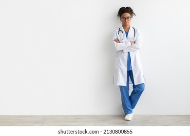 Serious Confident Female Black Doctor In Glasses Posing With Folded Arms Looking At Camera Standing Leaning On Wall, Lady Wearing Coat And Stethoscope, Full Body Length, White Studio Background Mockup