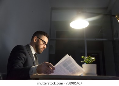 Serious Confident Male Employer Holding Business Note And Analyzing Advantages Of New Project At Night Shift In Dark Office Under Light Of Lamp