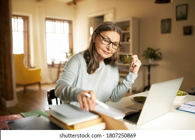 Serious confident middle aged female journalist with cup of coffee sitting at desk with books and laptop, making research, studying literature. Attractive mature woman copywriter working from home - Shutterstock ID 1807250869