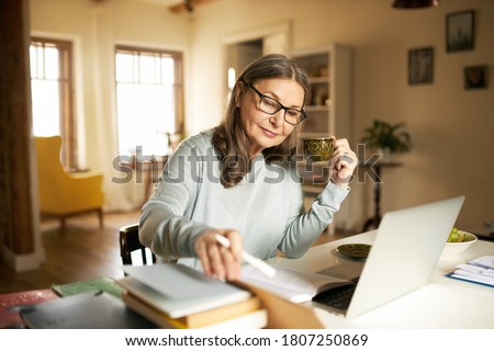 Serious confident middle aged female journalist with cup of coffee sitting at desk with books and laptop, making research, studying literature. Attractive mature woman copywriter working from home