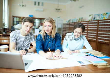 Serious confident student friends examining blueprint in library: concentrated guy pointing at laptop screen and explaining rules to groupmate