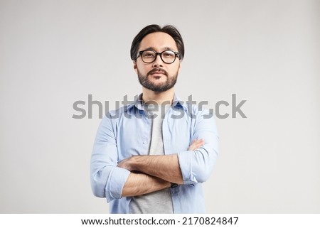 Serious confident young Asian web designer with black beard standing against isolated background and looking at camera