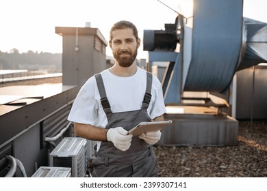 Serious construction worker in overalls and gloves standing and using modern tablet outdoors. Caucasian professional man surfing internet while working with devices on roof of factory.