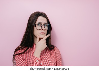 Serious contemplative woman holds chin, looks with thoughtful expression, feels doubt, raises eyebrows, dressed in shirt, isolated over pink background. People and emotion concept