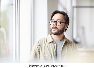 Serious contemplative young Asian man in casual shirt and eyeglasses sitting in office and looking out window - Powered by Shutterstock