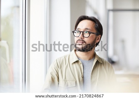 Serious contemplative young Asian man in casual shirt and eyeglasses sitting in office and looking out window