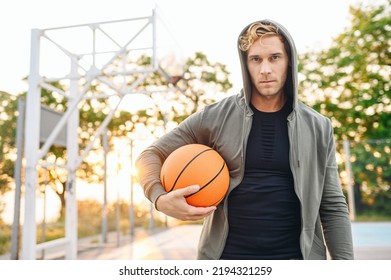 Serious Cool Confident Sunlit Young Sporty Sportsman Man In Grey Sports Clothes Hood Look Camera Training Hold In Hand Ball Play At Basketball Game Playground Court. Outdoor Courtyard Sport Concept.