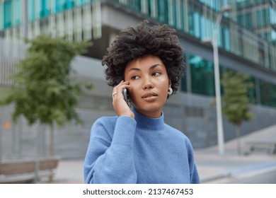 Serious Curly Haired Brunette Woman Has Telephone Conversation Wears Casual Blue Jumper Strolls Outdoors Poses Against Blurred Background. People Modern Technologies And Communication Concept