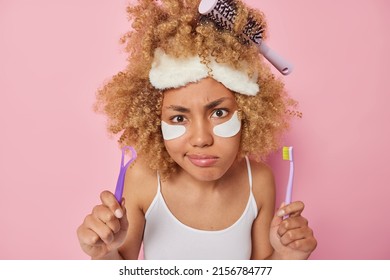 Serious curly haired young woman holds tooth brush and tongue scraper takes care of oral hygiene applies beauty patches under eyes to remove wrinkles wears headband has haircomb stuck in curls