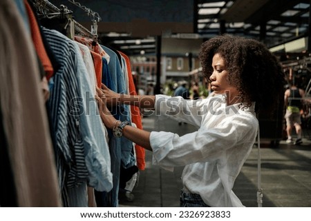Serious curly model choosing shirts in inside flea market. There are more stalls out of focus and some people over there.