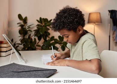 Serious curly-haired black boy sitting at desk and making notes in workbook during online lesson at home