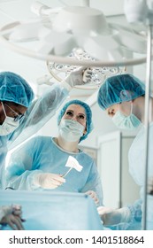 Serious Cute Female Doctor Holding Scissors With Bandage And Looking At The Lamp While Working With Colleagues