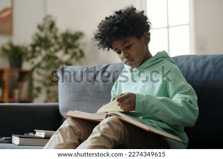 Serious cute schoolboy making notes in copybook while sitting on couch and reading article in encyclopaedia during preparation of homework