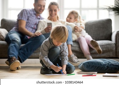 Serious cute son holding color marker drawing floor while parents and sister sitting apart  preschool kid loner boy playing alone  family leisure at home   creative child development concept