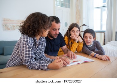 Serious Dad Sitting At Table With Kids And Reading Book. Bearded Father Explaining Task. Focused Girl And Interested Boys Looking At Book. Home Education, Family Time Concept