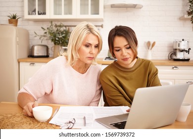 Serious dark haired girl sitting at kitchen table with her middle aged mother using wifi on generic portrable computer, drinking coffee and calculating family budget, planning expensive purchase - Powered by Shutterstock