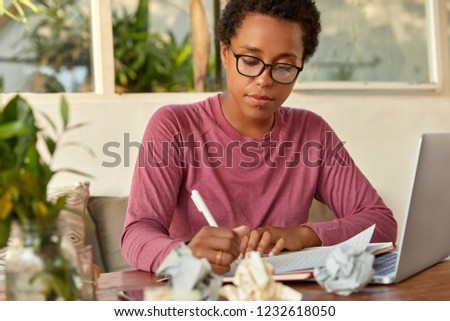 Serious dark skinned woman copywriter writes in sheet of paper, watches tutorial video on laptop computer, wears spectacles, models against cozy interior, scraps on table. People, technology, work