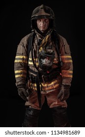 Serious Dirty Man In Uniform Of Firefighter And Helmet Standing On Black Background Looking Seriously At Camera. 