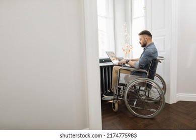 Serious Disabled Young Man Using Laptop For Remote Job At Home. Confident Caucasian Male In Wheelchair Surfing Internet Near Window At Workplace.