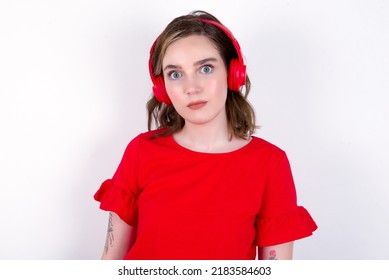 Serious Displeased Young Caucasian Woman Wearing Red T-shirt Over White Background Looks Puzzled At Camera Being Angry Wears Stereo Headphones Listens Music While Walking At Street